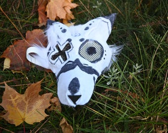 May include: A white furry mask with black painted details, including a cross, an eye with a mesh covering, and a nose. The mask is lying on a bed of green grass and brown leaves.