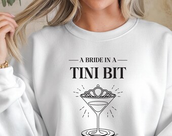 May include: White sweatshirt with the text "A Bride in a Tini Bit" above a martini glass design. The martini glass has a tiara on top and is surrounded by radiating lines. A casual, comfortable top for a bachelorette party or bridal event.