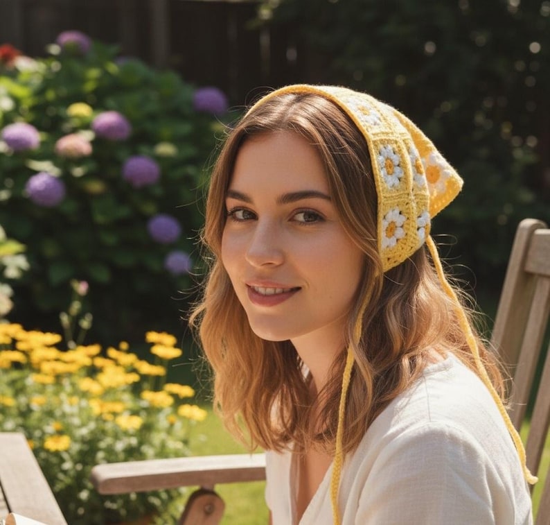 May include: A yellow crochet headscarf with white daisy accents. The headscarf is tied at the back and is worn by a person. The background features a garden with flowers and a wooden chair.
