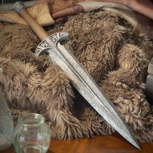 May include: A silver-colored dagger with a wooden handle. The blade has a textured surface and a pointed tip. The hilt features an ornate design. The dagger is displayed on a brown, furry surface.