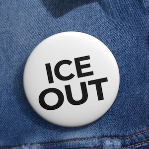 以下が含まれることがあります： 「ICE OUT」の文字が黒い太字の大文字で書かれた白いボタンバッジ。青いデニムジャケットの上に配置されています。