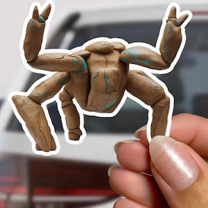 Puede incluir: Una figura marrón con forma de cangrejo con una textura agrietada y detalles turquesas. La figura tiene múltiples patas y brazos, con los brazos levantados en un gesto. La figura está sujeta por una mano.