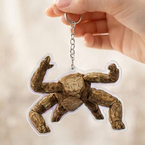 May include: A brown, textured keychain featuring a stylized figure with raised arms and legs. The figure has a rough, stone-like appearance and is attached to a silver keyring and chain. The keychain is held against a soft, blurred background.