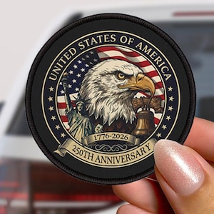May include: A circular black patch with a design celebrating the United States' 250th anniversary. It features a bald eagle, the Statue of Liberty, and the Liberty Bell, with text reading "UNITED STATES OF AMERICA" and "1776-2026".