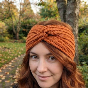 May include: A burnt orange crochet headband featuring a ribbed texture and a central twisted knot detail. The accessory is crafted with a consistent knit pattern that wraps around the head, providing a wide band coverage with a warm tone.