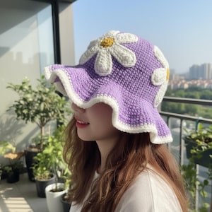 May include: A lavender crochet bucket hat featuring large white daisy appliqués with yellow centres. The hat has a wide, wavy brim edged in white yarn, constructed with a dense, textured stitch pattern throughout the crown and sides.