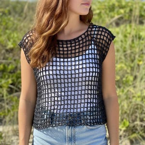 May include: A black crochet top with a square net design. The top is sleeveless and has a round neckline. A white tank top is visible underneath. The top is paired with blue jeans.