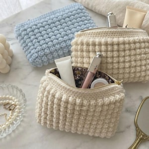 May include: Three rectangular crochet pouches with a textured bobble stitch pattern. One pouch is light blue, and two are cream-colored. The open cream pouch reveals a dark floral fabric lining and contains a white tube, a silver lipstick, and a compact.