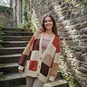May include: A long-sleeve, button-front cardigan featuring a patchwork design of square panels in shades of cream, tan, rust orange, and dark brown. The knit garment has a V-neckline, ribbed cuffs, and a ribbed hem, constructed with a textured stitch.