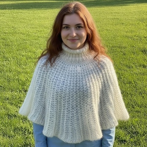 Può includere: Un poncho lavorato a maglia color crema con un alto collo a lupetto e una trama a coste su tutta la superficie. Il capo presenta una silhouette svasata a mantella che ricade sulle spalle e sul busto, realizzato in filato morbido e soffice.