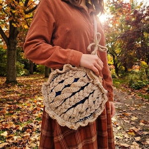 Könnte beinhalten: Eine runde, beige Häkeltasche mit strukturiertem Zopfmuster und gerüschtem Rand. Die Tasche hat einen langen Riemen mit einer dekorativen Schleife und zeigt ein dunkelblaues Innenfutter. Das Design erinnert an einen Kuchen mit Gitterkruste.
