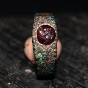 Puede incluir: Un anillo antiguo con una piedra preciosa de color rojo oscuro engastada en una banda desgastada de color bronce. La piedra preciosa presenta un diseño grabado. El anillo muestra signos de edad y desgaste, con una pátina verde visible en el metal.