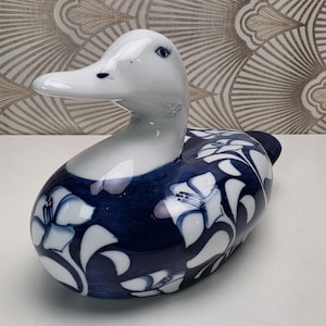 Op de afbeelding: Een keramieken eendenbeeldje met een glanzende witte kop en nek, voorzien van geschilderde blauwe ogen en wimpers. Het lichaam is versierd met een diepblauwe achtergrond en witte bloemmotieven, met een glad en reflecterend oppervlak.