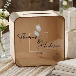 Op de afbeelding: Een houten trouwkaartendoos met de namen "Thomas & Madison" en de datum "10.12.24" op de voorkant. De doos is omgeven door huwelijksdecoratie, waaronder een "Cadeaus & Kaarten" bord, bloemen en een kaars.