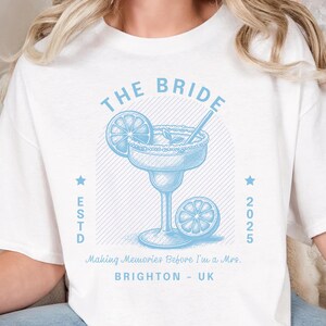 Może przedstawiać: Biała koszulka z jasnoniebieską grafiką margarity. Projekt zawiera napisy "THE BRIDE" i "ESTD 2025" oraz frazę "Making Memories Before I'm a Mrs. BRIGHTON - UK".