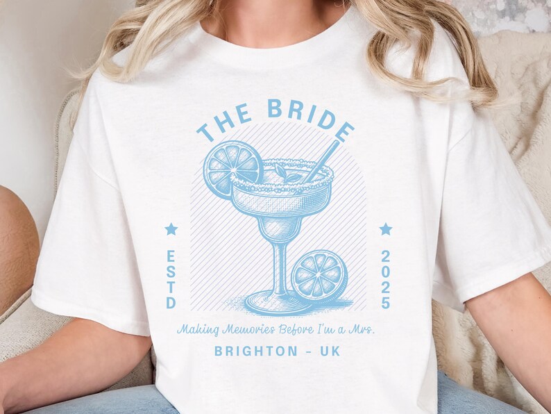 Może przedstawiać: Biała koszulka z jasnoniebieską grafiką margarity. Projekt zawiera napisy "THE BRIDE" i "ESTD 2025" oraz frazę "Making Memories Before I'm a Mrs. BRIGHTON - UK".