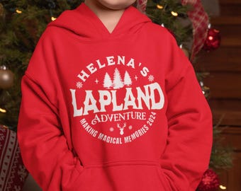 Può includere: Una felpa con cappuccio rossa con una grafica circolare bianca che recita "Helena's Lapland Adventure Making Magical Memories 2024". Il design include alberi, fiocchi di neve e una renna. La felpa ha una tasca frontale.