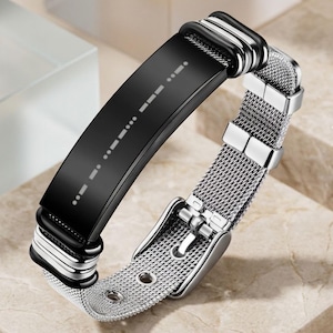 Puede incluir: Una pulsera que presenta una placa central rectangular negra, lisa y curva, con un patrón de línea punteada blanca. La banda está hecha de un material de malla metálica plateada e incluye un mecanismo de cierre de hebilla con metal.