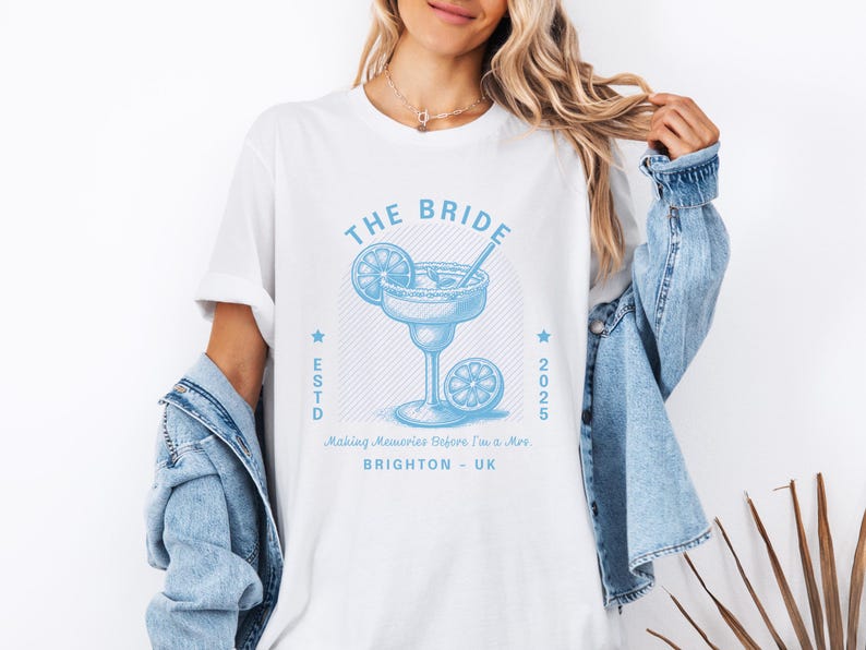 Może przedstawiać: Biała koszulka z jasnoniebieską grafiką margarity i napisem "THE BRIDE". Projekt zawiera koktajl, plasterki cytryny oraz napis "ESTD 2025 BRIGHTON - UK".