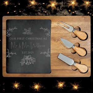 Może przedstawiać: Drewniana deska do serów z czarną wkładką z łupków i trzema nożami do sera. Na łupku wygrawerowano napis "OUR FIRST CHRISTMAS AS Mr. & Mrs. Williams EST 2025" oraz motywy kwiatowe. Noże mają drewniane uchwyty.