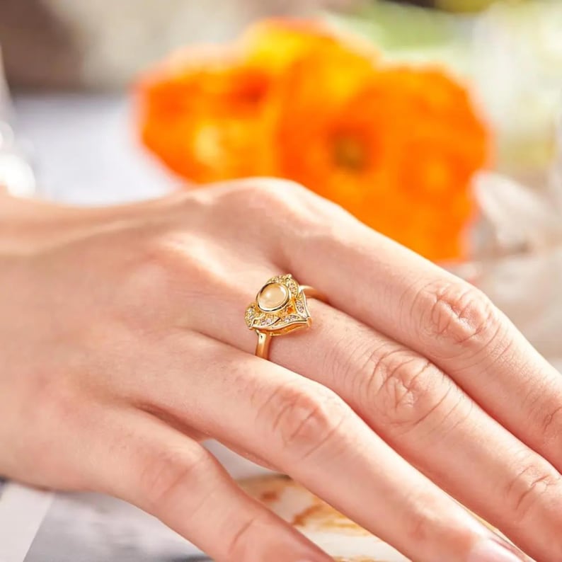 Puede incluir: Un anillo de oro con un dise&ntilde;o en forma de coraz&oacute;n. El anillo presenta una piedra central de color claro rodeada de piedras m&aacute;s peque&ntilde;as y transparentes. El anillo est&aacute; en un dedo, con flores naranjas en el fondo.