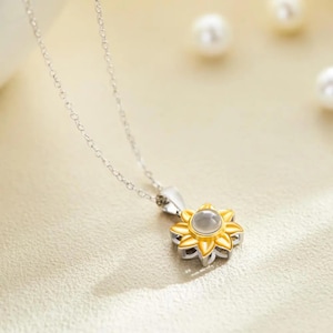 Puede incluir: Un collar de cadena plateada con un colgante de girasol. El colgante tiene una flor dorada con un centro claro y redondo. El collar se muestra sobre una superficie clara con perlas borrosas en el fondo.