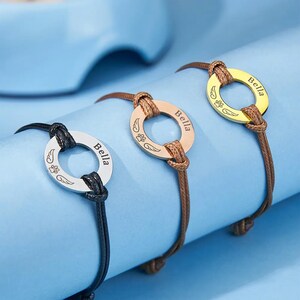 May include: Three personalised pet memorial bracelets with a round charm engraved with the name "Bella" and a paw print with wings. The bracelets are made with brown, black, and gold cord.