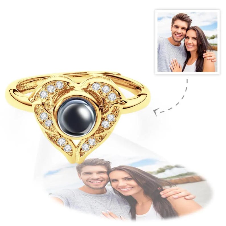 Puede incluir: Un anillo dorado con dise&ntilde;o de coraz&oacute;n. El anillo presenta una piedra oscura central rodeada de peque&ntilde;as piedras preciosas transparentes. La forma de coraz&oacute;n est&aacute; adornada con detalles decorativos. Una peque&ntilde;a foto de una pareja est&aacute; en el fondo.