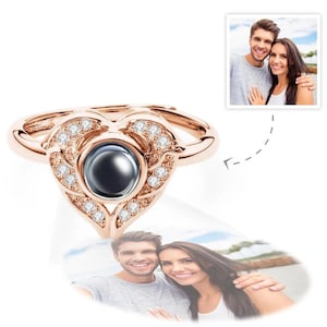 Puede incluir: Anillo en forma de coraz&oacute;n de oro rosa con una piedra oscura central y cristales claros circundantes. El anillo presenta un dise&ntilde;o &uacute;nico con una funci&oacute;n de proyecci&oacute;n. La imagen tambi&eacute;n incluye dos fotos de una pareja.