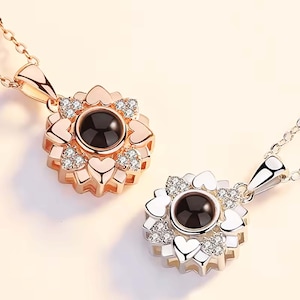 May include: Two silver necklaces with a projection pendant in the shape of a flower with hearts around the centre. The pendant on the left is rose gold and the pendant on the right is silver.