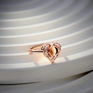 Puede incluir: Anillo de oro rosa con dise&ntilde;o de coraz&oacute;n. El anillo presenta una piedra central ovalada de color &aacute;mbar, rodeada de peque&ntilde;as piedras preciosas transparentes. El coraz&oacute;n est&aacute; detallado con delicada filigrana y est&aacute; engastado en una banda fina.