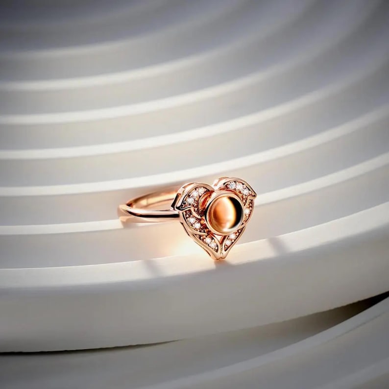 Puede incluir: Anillo de oro rosa con dise&ntilde;o de coraz&oacute;n. El anillo presenta una piedra central ovalada de color &aacute;mbar, rodeada de peque&ntilde;as piedras preciosas transparentes. El coraz&oacute;n est&aacute; detallado con delicada filigrana y est&aacute; engastado en una banda fina.