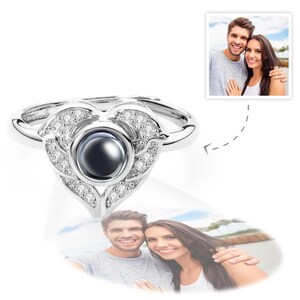 Puede incluir: Un anillo plateado con un dise&ntilde;o en forma de coraz&oacute;n. El anillo presenta una piedra oscura central rodeada de peque&ntilde;as piedras claras. La forma de coraz&oacute;n est&aacute; adornada con elementos decorativos. El anillo se muestra con la foto de una pareja.