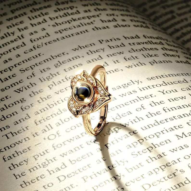 Puede incluir: Un anillo dorado con una piedra oscura central, rodeado por un dise&ntilde;o floral y peque&ntilde;as gemas transparentes. El anillo se presenta sobre un libro abierto, creando una est&eacute;tica cl&aacute;sica y elegante.