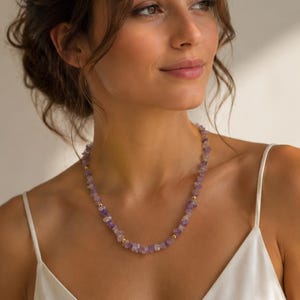 May include: A delicate necklace featuring small, irregular amethyst beads interspersed with gold-colored beads. The necklace is worn against a white camisole, creating a contrast that highlights the necklace's purple hues. The jewelry is a simple yet elegant accessory.