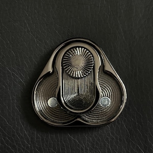 May include: A close-up of a silver decorative clasp. The clasp features a distinctive design with a central sunburst element and two circular, ridged sections. The background is a textured, dark grey surface.