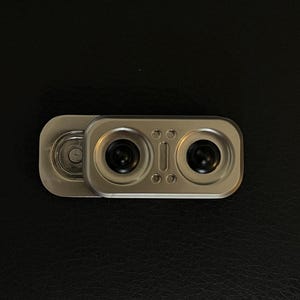 May include: A rectangular silver-toned metal sliding webcam cover featuring two circular black lens openings. The device has a smooth, brushed metallic finish with a small vertical indentation between the two circular cutouts on the sliding panel.