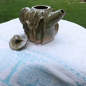 Puede incluir: Una tetera de cerámica verde con un diseño texturizado y orgánico. La tetera tiene una tapa con una manija y un pico. La tetera está sentada sobre un mantel blanco con un patrón floral azul.