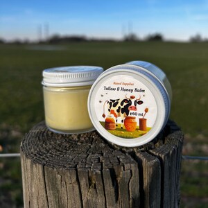 May include: A white jar with a yellow balm inside. The jar has a white lid with a colorful label that says "Based Supplies Tallow & Honey Balm" and features a cartoon cow and honeycombs. The label also says "60 ml".