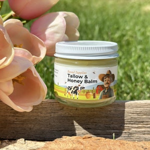 Puede incluir: Un pequeño frasco de vidrio con tapa blanca lleno de un bálsamo amarillo. La etiqueta del frasco dice "Based Supplies Tallow & Honey Balm" y presenta una vaca, una colmena y un granjero de dibujos animados.