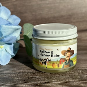 May include: A white jar with a yellow label that says "Based Supplies Tallow & Honey Balm". The label features a cartoon cow, a bee, and a man in a straw hat.