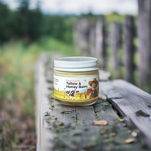May include: A glass jar of Tallow & Honey Balm by Based Supplies. The jar has a white lid and a colorful label with a cartoon illustration of a farmer, a cow, and a beehive.