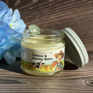 May include: A glass jar with a white lid containing a yellow balm. The label on the jar reads "Based Supplies Tallow & Honey Balm" and features a cartoon illustration of a farmer, a cow, and a beehive.
