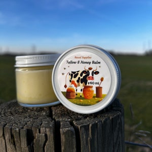 May include: A white jar with a silver lid containing a yellow balm. The lid has a colorful label with a cartoon cow, honeycombs, and the text "Based Supplies Tallow & Honey Balm 60 ml".