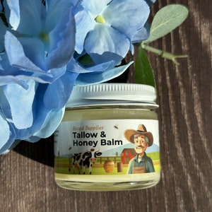 Puede incluir: Un pequeño frasco de vidrio con tapa blanca lleno de un bálsamo amarillo. La etiqueta del frasco dice "Based Supplies Tallow & Honey Balm" y presenta una ilustración de dibujos animados de una vaca, una abeja, un granjero y un tarro de miel.