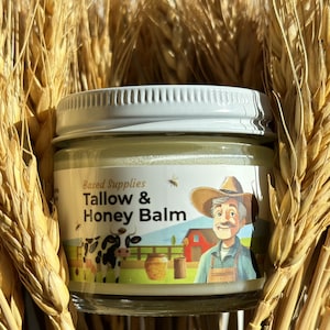 Puede incluir: Un pequeño frasco de vidrio con tapa blanca que contiene un bálsamo amarillo. La etiqueta del frasco presenta una ilustración de dibujos animados de un granjero, una vaca, una colmena y un granero rojo. La etiqueta dice "Based Supplies Tallow & Honey Balm".