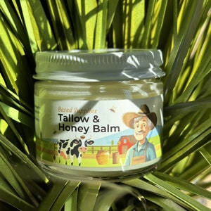 Op de afbeelding: Een klein glazen potje met een wit deksel dat een gele balsem bevat. Het etiket vermeldt "Based Supplies Tallow & Honey Balm" en toont een cartoonillustratie van een boer, een koe en een honingraat.