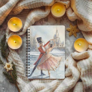 Puede incluir: Un cuaderno con espiral con una escena invernal que representa un Cascanueces y una bailarina bailando frente a un castillo. El cuaderno está rodeado de velas encendidas, una estrella decorativa y una manta de punto color crema.