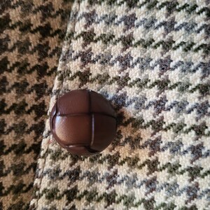 May include: Close-up of a brown leather button on a houndstooth patterned fabric. The fabric features a repeating pattern in shades of brown, gray, and cream. The button is round and has a textured surface.