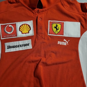 May include: Red polo shirt with a collar and button-up placket. Features embroidered patches including the Vodafone logo, Shell logo, Ferrari emblem, Bridgestone, and Puma logo. The shirt has a white panel on the side.
